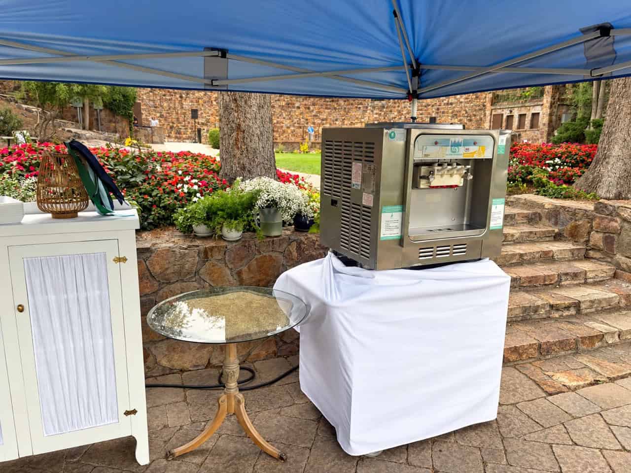 Soft serve machine set up under a canopy at the American Fork Amphitheater, with the stone wall and flower beds behind it
