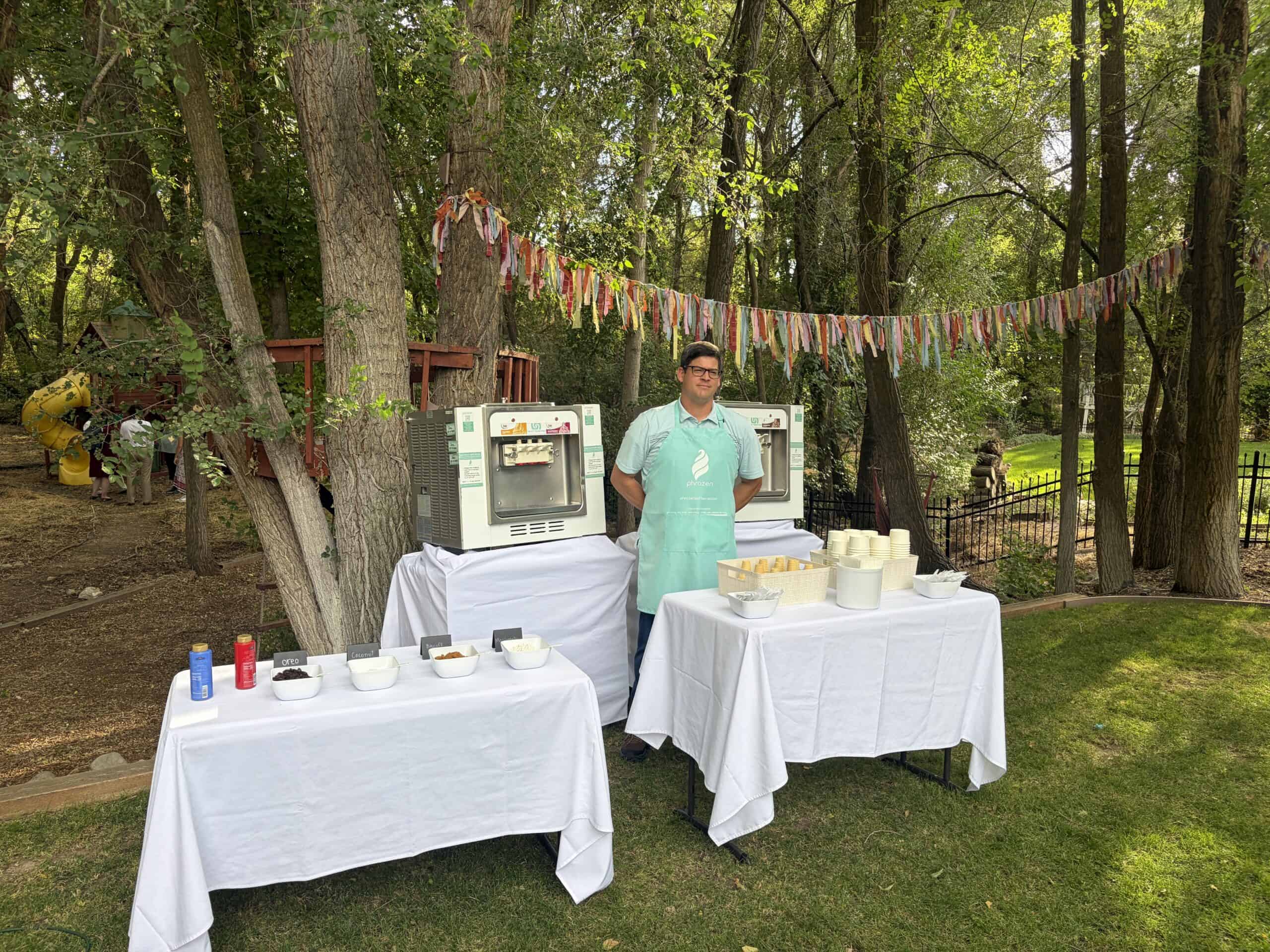 A Phrozen Soft Serve server in a teal apron beside two machines and a full toppings table at a backyard wedding in Provo