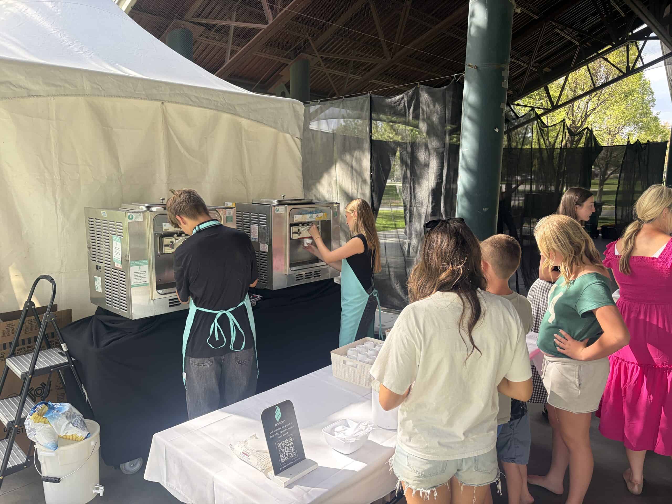 Two Phrozen Soft Serve staff in teal aprons serving a line of guests at a company party at Electric Park in Lehi